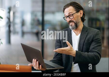 Glücklicher leitender Manager, der mit Kollegen über den Arbeitsprozess spricht. Erfolgreicher Geschäftsmann mit Laptop im modernen Büro, lächelnd Stockfoto