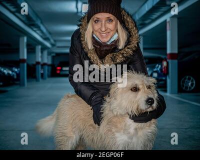 Blonde kaukasische Frau trägt Winterkleidung, posiert glücklich mit ihrem Schäferhund in einer Parkgarage Stockfoto
