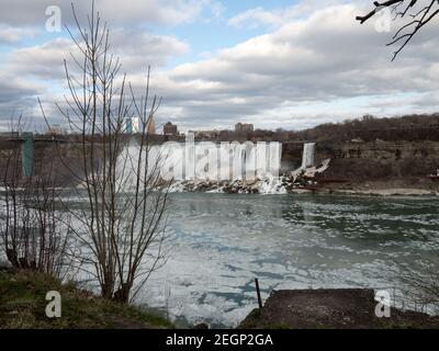 Niagarafälle amerikanische Seite von Kanada aus gesehen, liegt Eis über dem Wasser und gefrorener Nebel am Boden der Fälle, blauer Himmel und Gebäude im Hintergrund Stockfoto