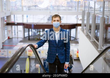 Junger Kaukasier in einer Jacke und einer medizinischen Maske Steigt die Rolltreppe in einem Einkaufszentrum Stockfoto