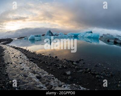 Sonnenuntergang über der Jokulsarlon Gletscherlagune in Island, goldene Farben spiegeln sich auf dem Wasser und kontrastieren mit den blauen Eisbergen, im Vordergrund sind es Stockfoto