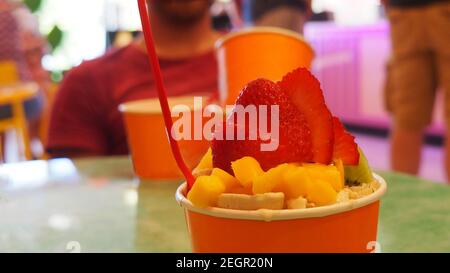 Eiscreme mit Fruchtauflagen in orangefarbener Tasse mit rotem Löffel, mehr Tassen und Menschen außerhalb des Fokus im Hintergrund Stockfoto