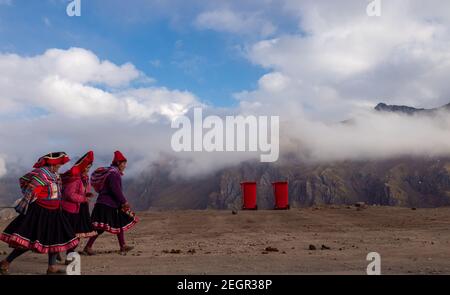 Peru, Vinicunca - 1. Oktober 2019 - drei indigene Frauen gehen vor der wolkenbedeckten Bergkette in traditioneller Kleidung, zwei rote Thrash b Stockfoto