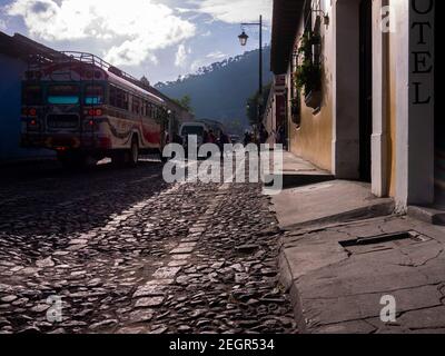 Guatemala, Antigua - 26. Mai 2019 - Schulkinder überqueren Straße vor bunten öffentlichen Bus, Straße ist Kopfsteinpflaster Stockfoto