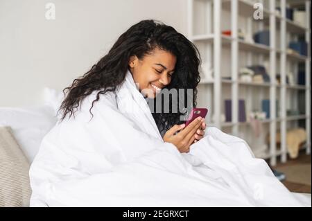 Glückliche junge afroamerikanische Frau mit lockigem Haar in einer gemütlichen weichen Decke zu Hause im warmen Bett, SMS-Chat mit Freunden auf Stockfoto