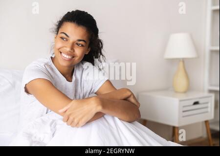 Positive junge entspannte Mixed-Race-Frau mit gemütlichen warmen weichen Decke bedeckt sitzen in ihrem Pyjama und zu Hause bleiben, Tagträumen, Ausruhen und Stockfoto