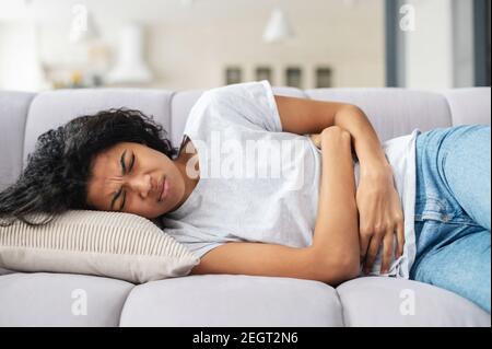 Traurige junge afroamerikanische Frau trägt Freizeitkleidung, die unter Menstruationsschmerzen leidet, sich krank zu ihrem Bauch fühlt, Bauch hält, Bauch hat Stockfoto