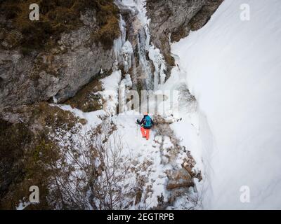 Luftdrohnenaufnahme eines Wanderers mit Rucksack, orangefarbenem Hut und orangefarbener Hose, die neben dem Wasserfall stehen, umgeben von Eiszapfen und Schnee im Winter Stockfoto