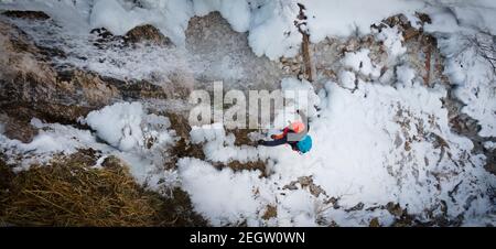 Luftdrohnenaufnahme eines Wanderers mit Rucksack, orangefarbenem Hut und orangefarbener Hose, die neben dem Wasserfall stehen, umgeben von Eiszapfen und Schnee im Winter Stockfoto