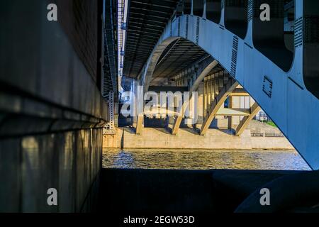 Leistungsstarke Eisenbrücke Struktur, Blick von unten. Betonsäulen, große eiserne Nieten. Unter der Brücke fließt ein Fluss. Perspektive. Stockfoto