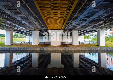 Leistungsstarke Eisenbrücke Struktur, Blick von unten. Betonsäulen, große eiserne Nieten. Unter der Brücke fließt ein Fluss. Perspektive. Stockfoto