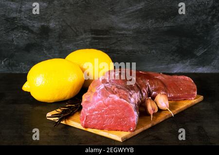 Frisches rohes Kalbsblatt in einem großen Stück mit Zitronen, Zweig Rosmarin und Knoblauch auf einem hölzernen Schneidebrett auf dunklem Vintage-Hintergrund. Die Konz Stockfoto