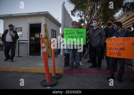 18. Februar 2021, Mexiko, Tijuana: Ein privater Sicherheitsbeamter, der den Grenzübergang bewacht, beobachtet eine Gruppe von Migranten, die Protestschilder tragen. Einer der Migranten (M) beschwert sich, dass er acht Monate auf einen Termin bei den US-Behörden gewartet habe. Zentralamerikanische und mexikanische Migranten, die Flüchtlinge im Flüchtlingsheim Agape sind, demonstrierten vor dem Haupteingang des Grenzübergangs El Chaparral an der Grenze zwischen Tijuana, Mexiko, Und San Diego, Kalifornien, mit dem Ziel, die US-Behörden aufzufordern, die Grenze zu öffnen und den Prozess der Gewährung von politischem Asyl für die Zuflucht wieder aufzunehmen Stockfoto