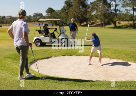 Vier kaukasische ältere Männer und Frauen spielen Golf und beobachten einen Mann, der einen Schuss aus dem Bunker vorbereitet Stockfoto
