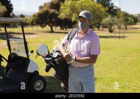 Kaukasischer älterer Mann mit Gesichtsmaske beim Spaziergang über den Golfplatz Golftasche in der Hand Stockfoto