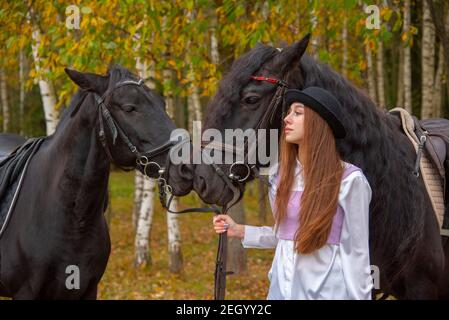 Ein rothaariges schlankes Mädchen steht in einem Herbsthain mit zwei Pferden. Stockfoto