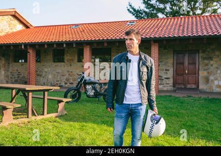 Junger Mann posiert im Freien mit benutzerdefinierten Motorrad im Hintergrund Stockfoto