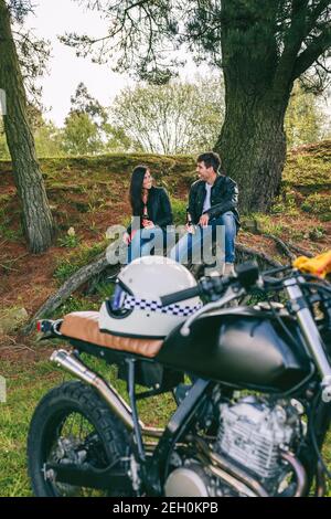 Junges Paar, das sich von einer Motorradtour ausruhte und ein Bier im Freien mit dem Motorrad im Vordergrund hatte. Selektiver Fokus auf Paar im Hintergrund Stockfoto