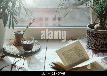 Orientalischer Kaffee in traditioneller türkischer Kupferkaffeekanne mit Blumen auf der Fensterbank. Rustikale Fensterbank aus Holz mit exotischen Pflanzen und altem Buch. Kalt Stockfoto