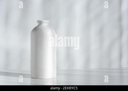 Minimalistisch elegante Komposition mit einer weißen Vase auf einem weißen Tisch. Helles Sonnenlicht und kontrastierende Schatten an einer weißen Wand. Stilvoller Minimalismus. Stockfoto