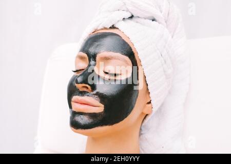 Schöne Frau mit Handtuch auf dem Kopf entspannend mit schwarzen reinigenden Kohle Gesichtsmaske. Kosmetologie und Hautpflege. Schönheitsbehandlungen. Spa-Therapie. Stockfoto