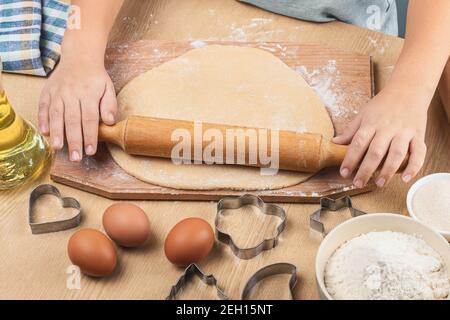 Kinderhände Rollen Teig für hausgemachte Kekse auf einem Holzbrett aus. Auf einem Holztisch gibt es ein blaukariertes Handtuch, Eier, ein Ausstechform, Butter Stockfoto