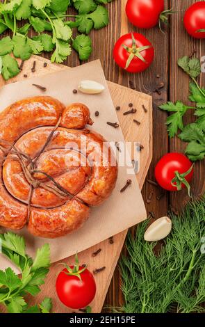 Hausgemachte köstliche, köstliche geräucherte Fleischwurst, mit Seil gebunden und in einer Spirale verdreht, liegt auf einem Holzbrett neben Dill, Petersilie, Tomaten Stockfoto