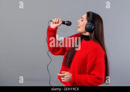 Junge, emotionale Brünette mit Kopfhörern und einem Mikrofon in einem roten Pullover gekleidet singt Karaoke, isoliert auf grau Stockfoto