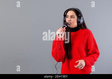 Junge, emotionale Brünette mit Kopfhörern und einem Mikrofon in einem roten Pullover gekleidet singt Karaoke, isoliert auf grau Stockfoto