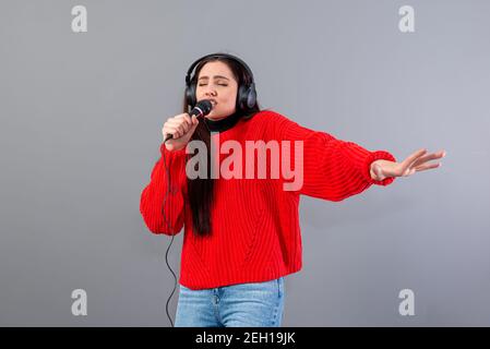 Junge, emotionale Brünette mit Kopfhörern und einem Mikrofon in einem roten Pullover gekleidet singt Karaoke, isoliert auf grau Stockfoto