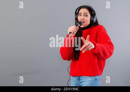 Junge, emotionale Brünette mit Kopfhörern und einem Mikrofon in einem roten Pullover gekleidet singt Karaoke, isoliert auf grau Stockfoto