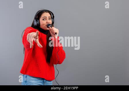 Junge, emotionale Brünette mit Kopfhörern und einem Mikrofon in einem roten Pullover gekleidet singt Karaoke, isoliert auf grau Stockfoto