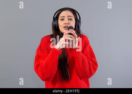 Junge, emotionale Brünette mit Kopfhörern und einem Mikrofon in einem roten Pullover gekleidet singt Karaoke, isoliert auf grau Stockfoto