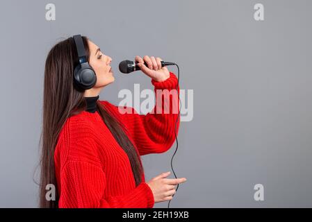 Junge, emotionale Brünette mit Kopfhörern und einem Mikrofon in einem roten Pullover gekleidet singt Karaoke, isoliert auf grau Stockfoto