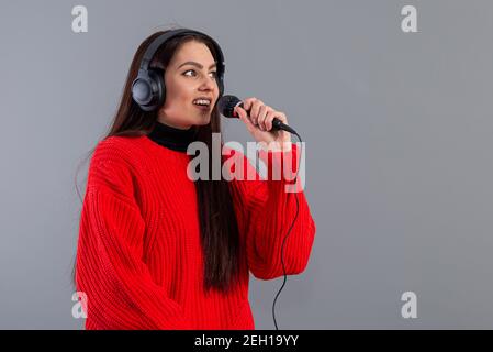 Junge, emotionale Brünette mit Kopfhörern und einem Mikrofon in einem roten Pullover gekleidet singt Karaoke, isoliert auf grau Stockfoto