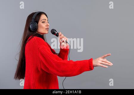 Junge, emotionale Brünette mit Kopfhörern und einem Mikrofon in einem roten Pullover gekleidet singt Karaoke, isoliert auf grau Stockfoto