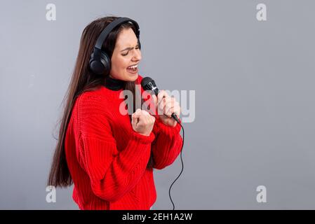 Junge, emotionale Brünette mit Kopfhörern und einem Mikrofon in einem roten Pullover gekleidet singt Karaoke, isoliert auf grau Stockfoto