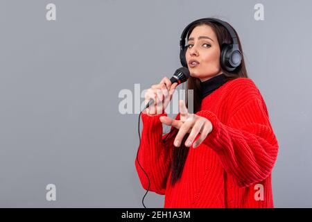 Junge, emotionale Brünette mit Kopfhörern und einem Mikrofon in einem roten Pullover gekleidet singt Karaoke, isoliert auf grau Stockfoto