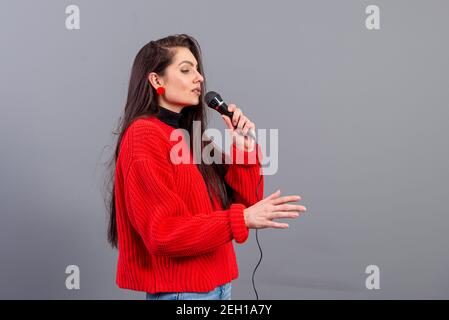 Junge, emotionale Brünette mit einem Mikrofon, das in einem roten Pullover gekleidet ist, singt Karaoke oder sagt eine Rede, isoliert auf Grau Stockfoto