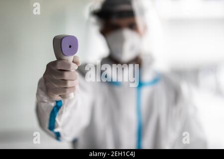 Mann Arzt in einem Schutzanzug aus dem Coronavirus, eine medizinische Maske Brille und Handschuhe, verwendet eine Infrarot-berührungslose Thermometer Pistole, um Körper te zu überprüfen Stockfoto