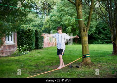 Ein Teenager-Junge balanciert auf einer schlaffen Linie, enge Seil, das zwischen zwei Bäumen im Garten gebunden ist. Ein Landhaus und eine Mauer sind im Hintergrund. Stockfoto