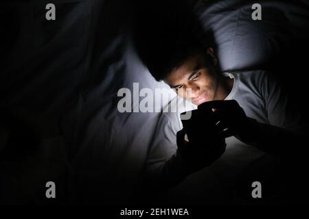 Junger Mann, der nachts mit einem Smartphone auf dem Bett sitzt Von oben nach unten Stockfoto