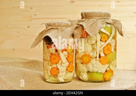 Konzept von zwei saisonalen Gurkenscheiben Glas mit farbigen grünen rohen Tomaten, orange Karotte und weißen Blumenkohl auf Holzhintergrund. Stockfoto