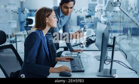 Moderne Fabrik Büro: Männliche Projektleiterin spricht mit einer weiblichen Industrieingenieurin, die auf Computer arbeitet. Professionelle Teamarbeit, Junge Spezialisten Stockfoto