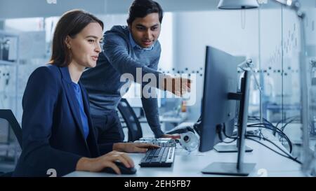 Moderne Fabrik Büro: Männliche Projektleiterin spricht mit einer weiblichen Industrieingenieurin, die auf Computer arbeitet. Professionelle Teamarbeit, Junge Spezialisten Stockfoto