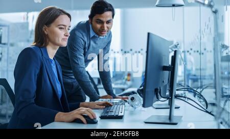 Moderne Fabrik Büro: Männliche Projektleiterin spricht mit einer weiblichen Industrieingenieurin, die auf Computer arbeitet. Professionelle Teamarbeit, Junge Spezialisten Stockfoto
