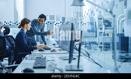 Inside Factory Office: Männliche Projektleiterin spricht mit einer weiblichen Industrieingenieurin, die am Computer arbeitet, sprechen. In Workshop: Professionelle Arbeiter Stockfoto