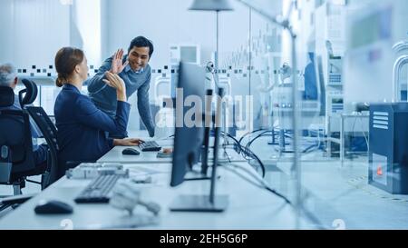 Inside Factory Office: Männliche Projektleiterin spricht mit einer weiblichen Industrieingenieurin, die am Computer arbeitet, geben Sie hoch fünf. In Workshop: Arbeiter Verwenden Stockfoto