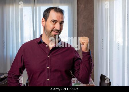 Attraktiver, lächelnder bärtiger Mann mit Zahnspangen in einem violetten Hemd, der seine Faust triumphierend hebt. Gewinner-Konzept Stockfoto