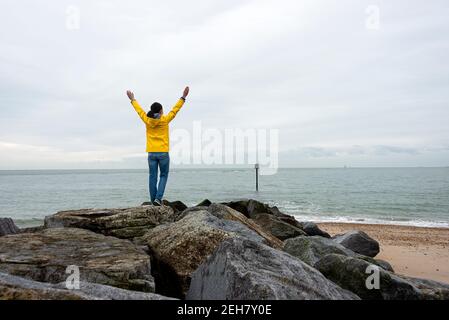 Rückansicht einer Frau, die an einem grau bewölkten Tag auf Felsen mit erhobenen Armen steht und auf das Meer blickt. Stockfoto